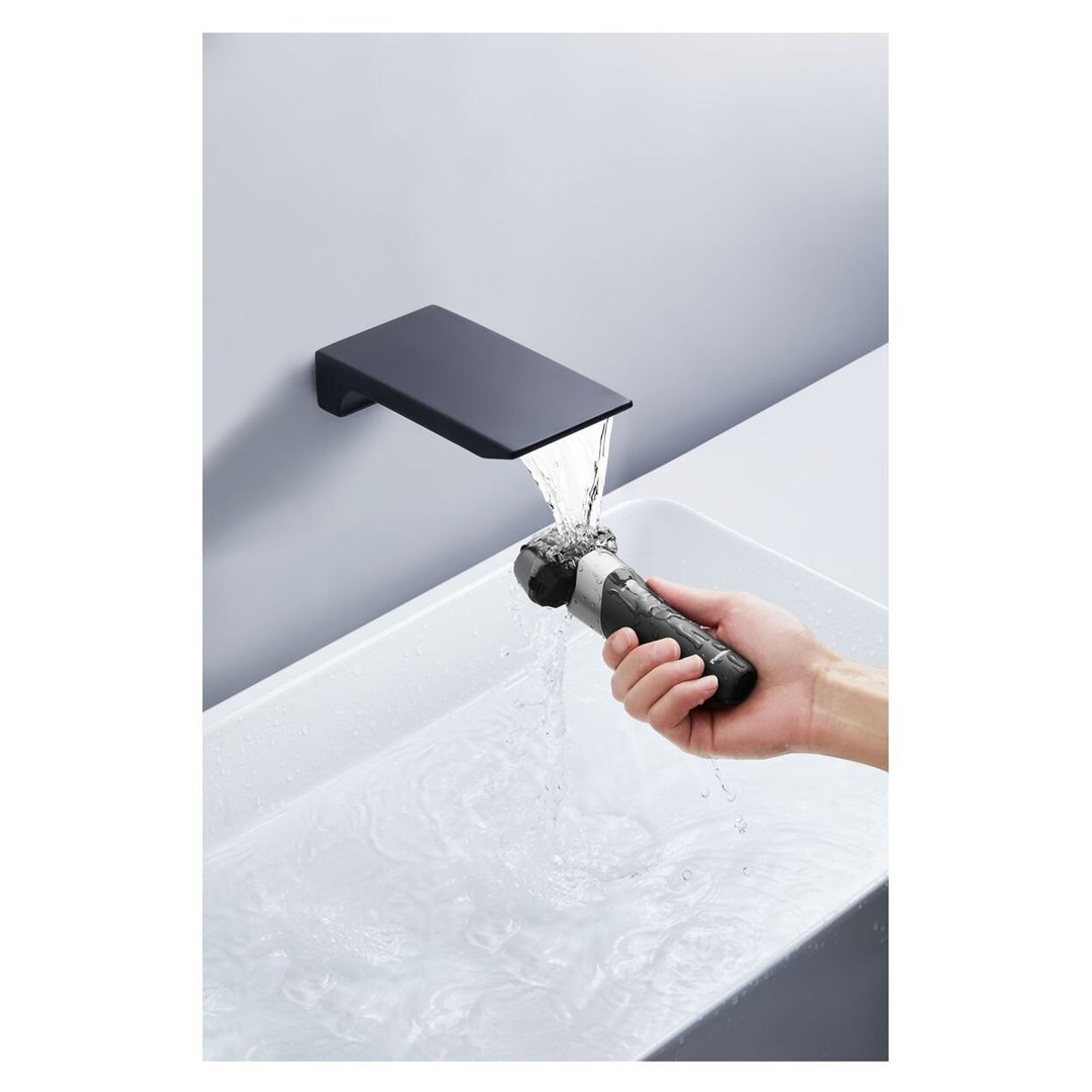 Hand holding a black electric shaver under running water from a wall-mounted faucet