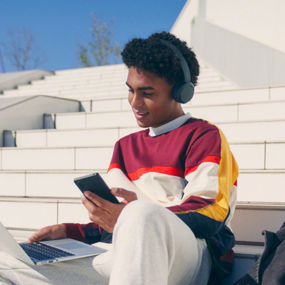 Young man wearing black Sony wireless headphones, using a laptop and phone outdoors