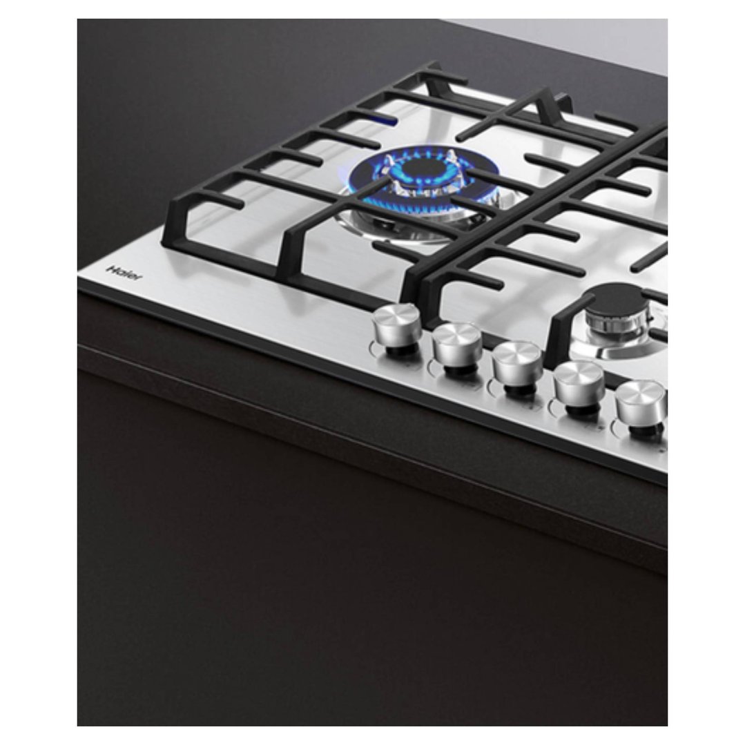 Stainless steel gas cooktop with five burners, one lit, on a dark countertop