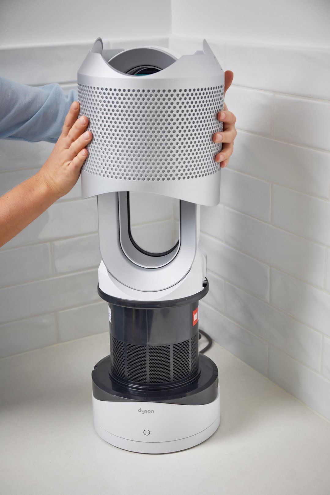 Hands removing the top section of a Dyson air purifier on a white counter