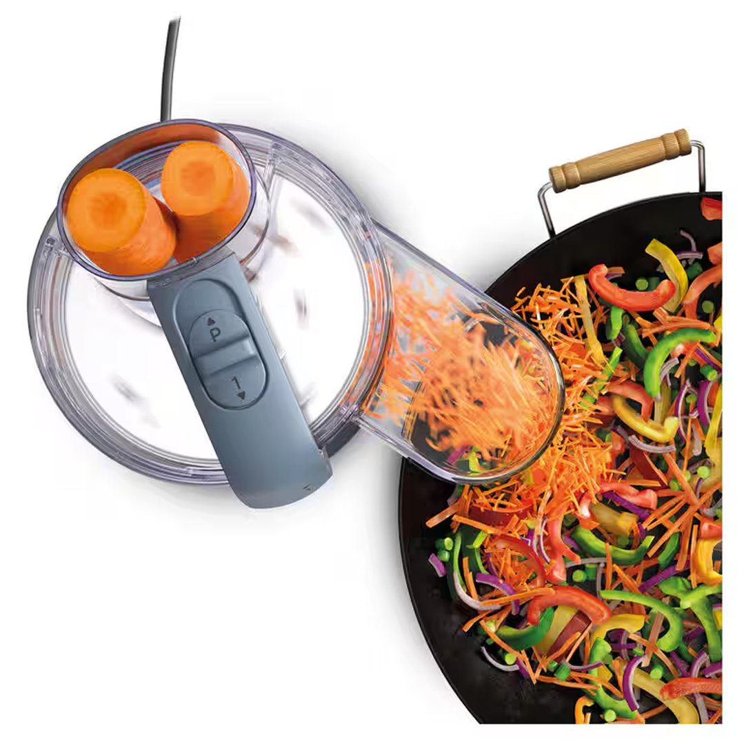 Food processor shredding carrots into a wok filled with colorful vegetables