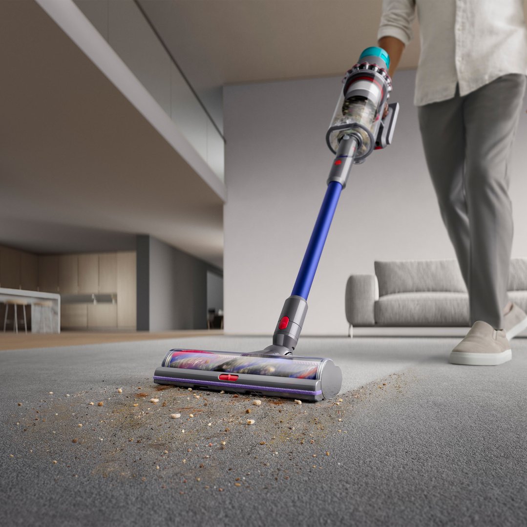 Person vacuuming a grey carpet with a Dyson handstick vacuum, showing dirt being collected