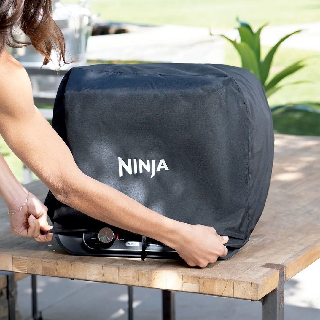 Person placing a black Ninja Woodfire Premium Outdoor Oven Cover on an oven