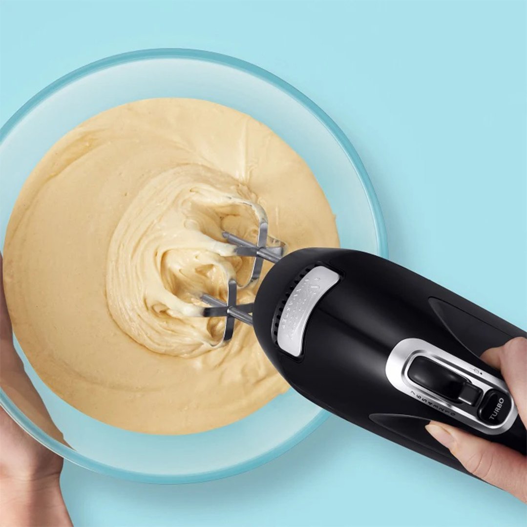 Hand mixer mixing batter in a glass bowl on a light blue background