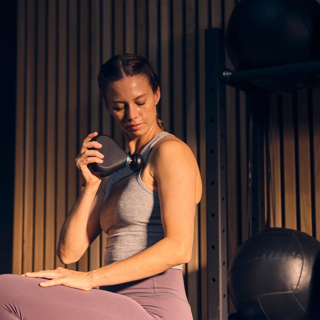 Woman using a Theragun Mini Plus massage gun on her shoulder in a gym setting