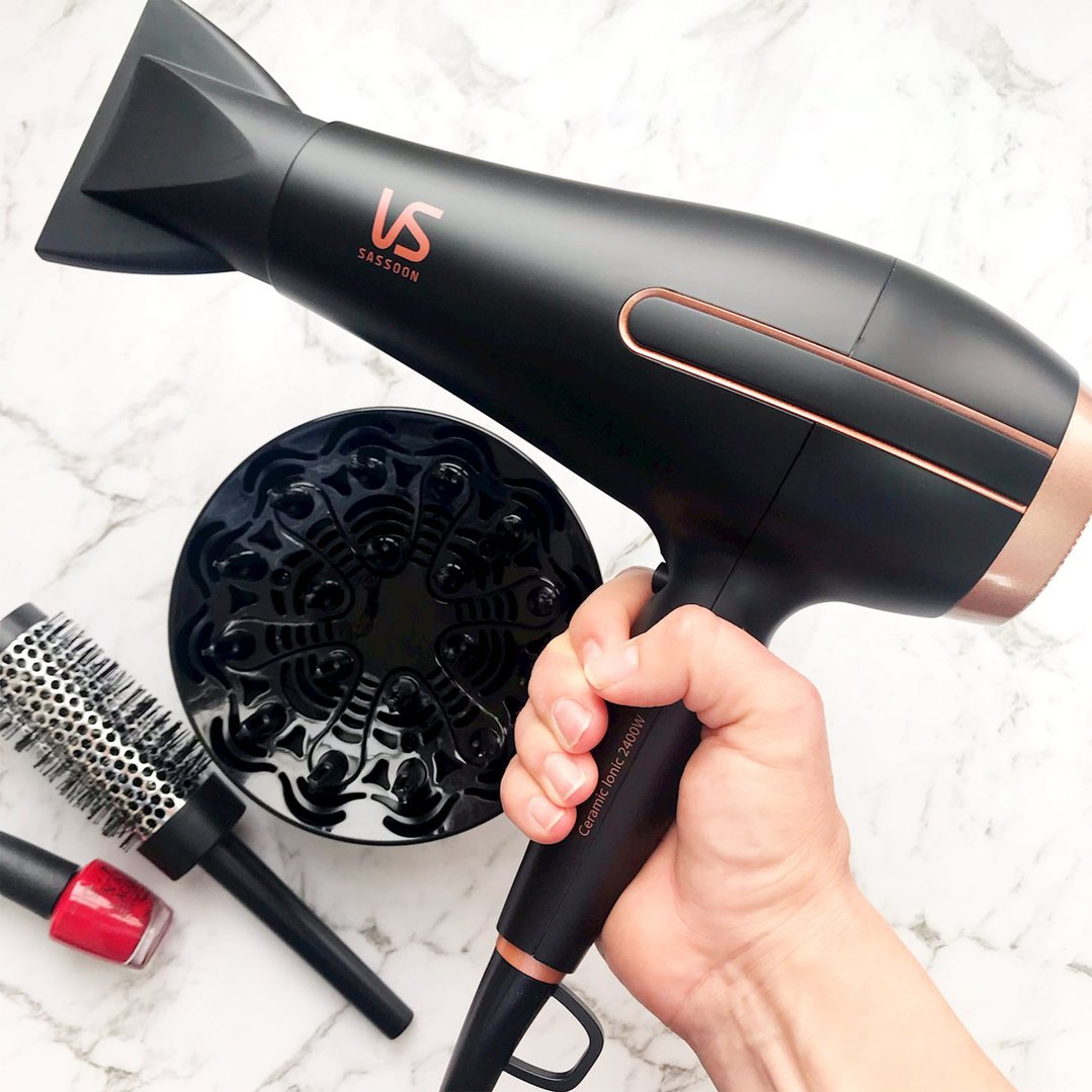Hand holding a black hair dryer with a diffuser, brush, and nail polish