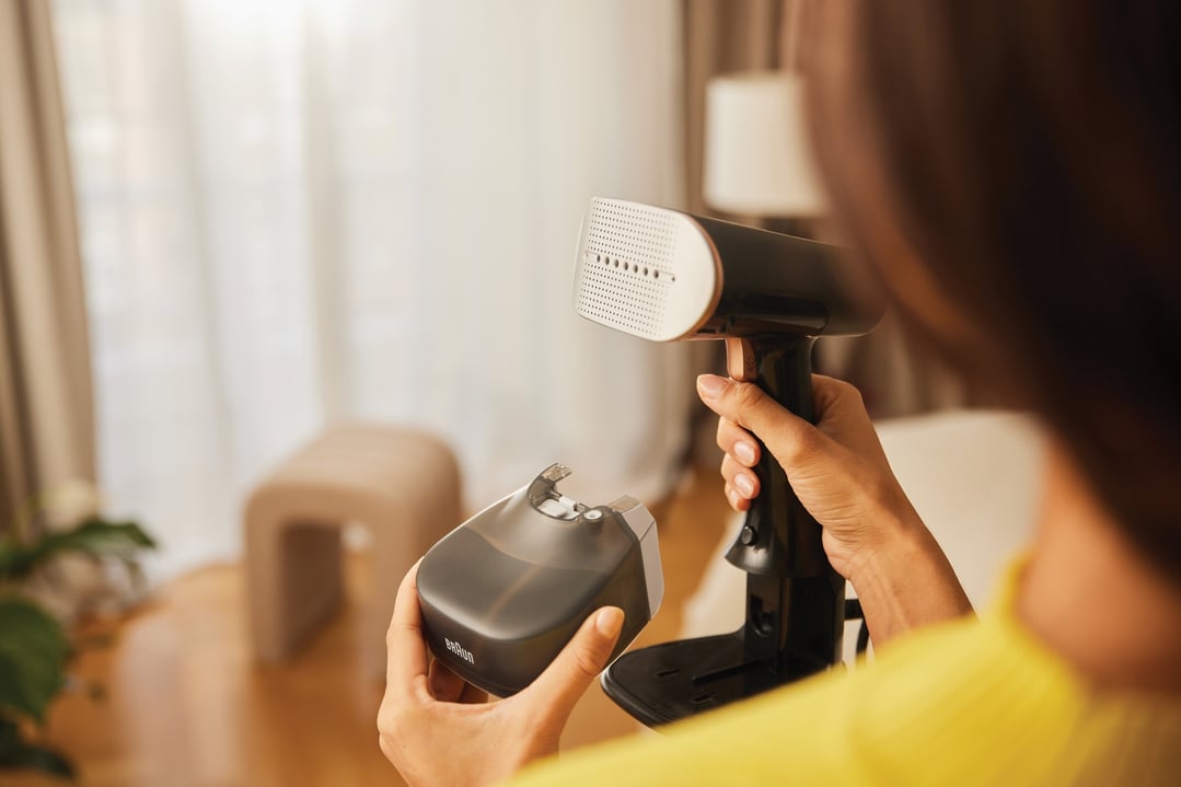 Person holding a Braun Garment Steamer and its water tank in a room