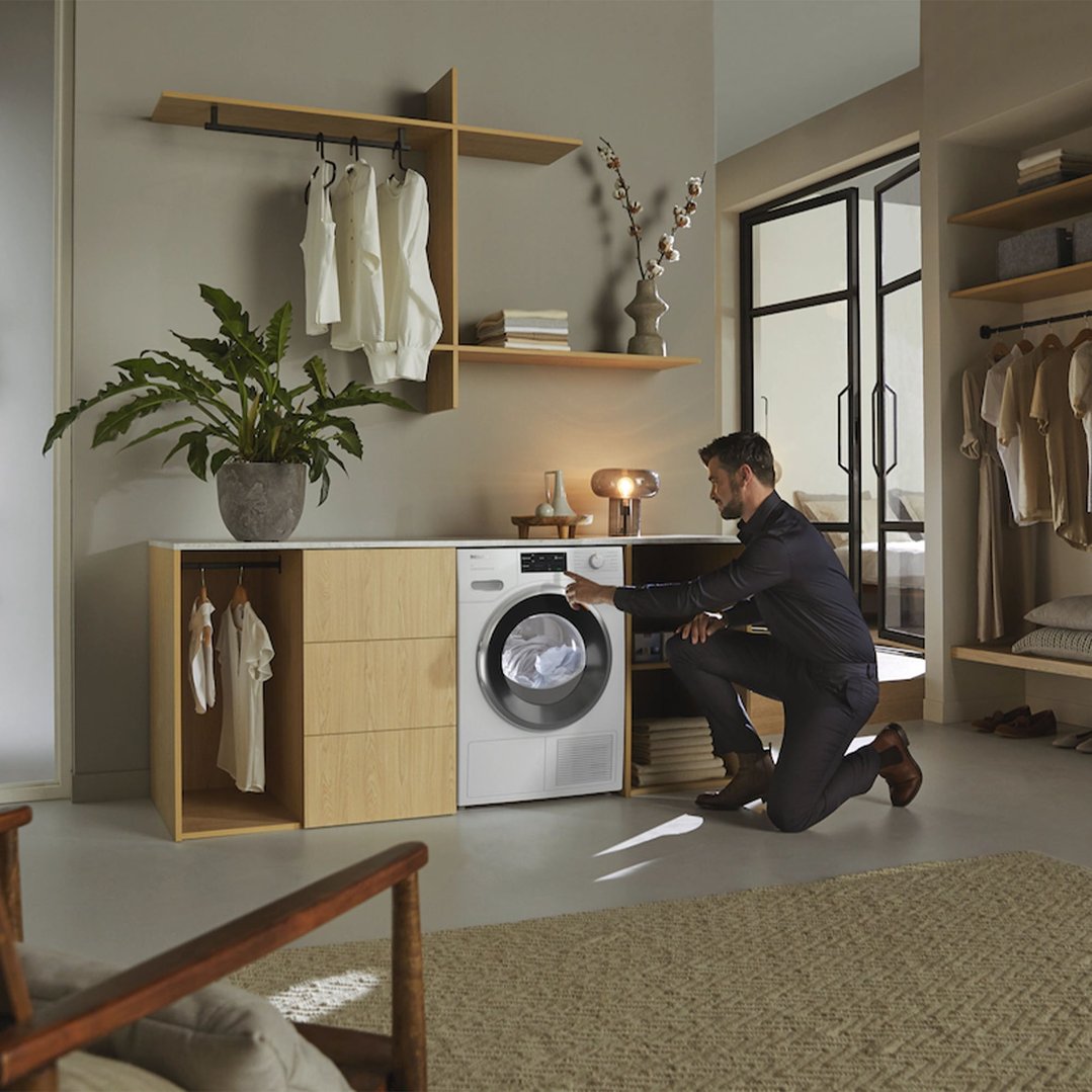 Man kneeling to operate a Miele heat pump dryer in a modern laundry room
