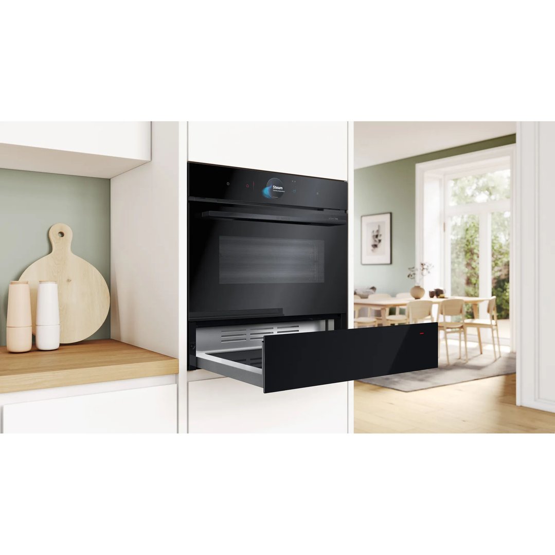 Black warming drawer and oven built into kitchen cabinetry with open plan dining area