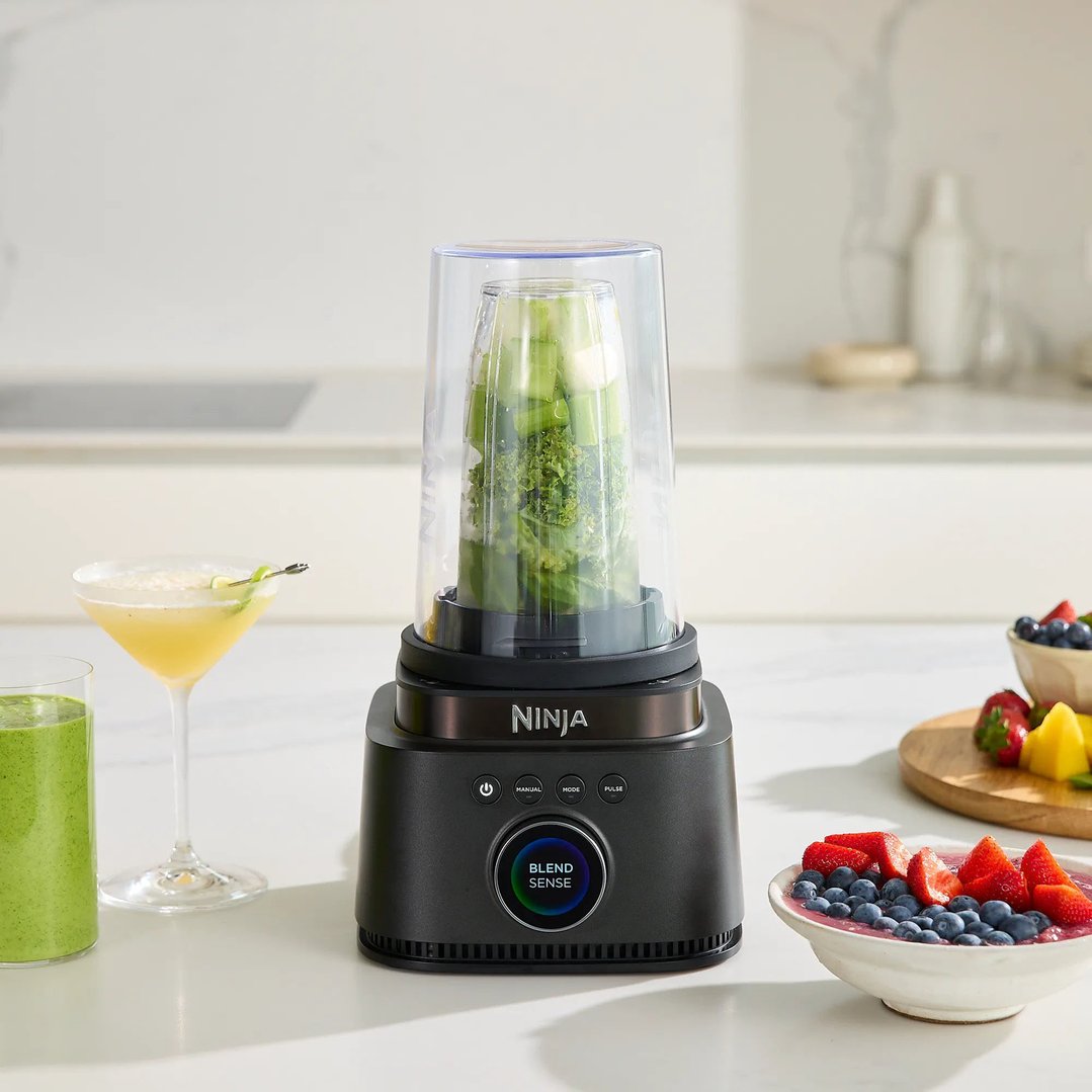 Ninja blender with green ingredients, drinks, and fruit on a white counter