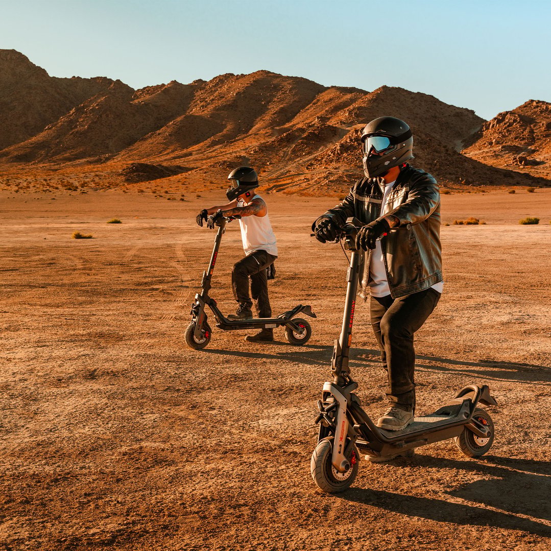 Two people on electric scooters in a desert landscape with mountains in the background