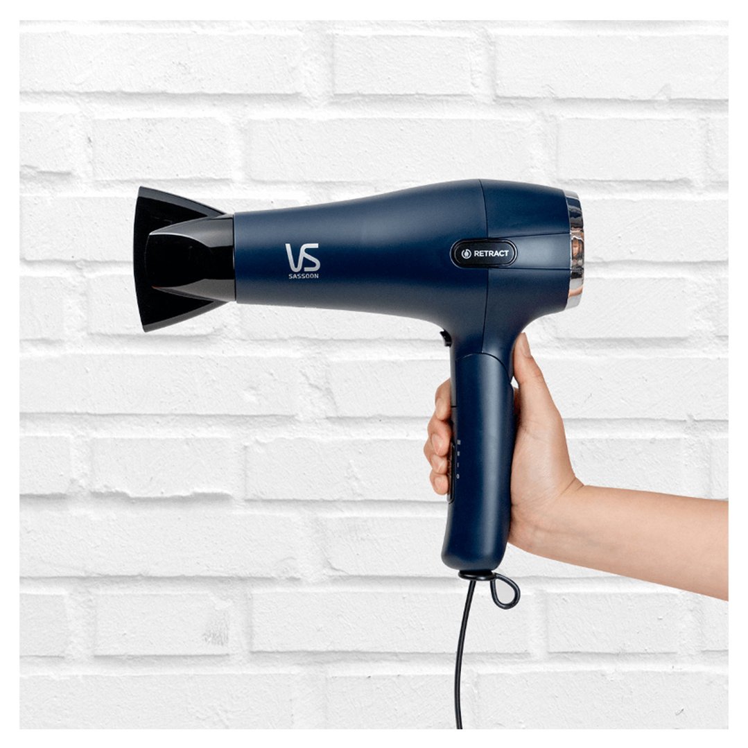 Hand holding a blue VS SASSOON hair dryer against a white brick wall