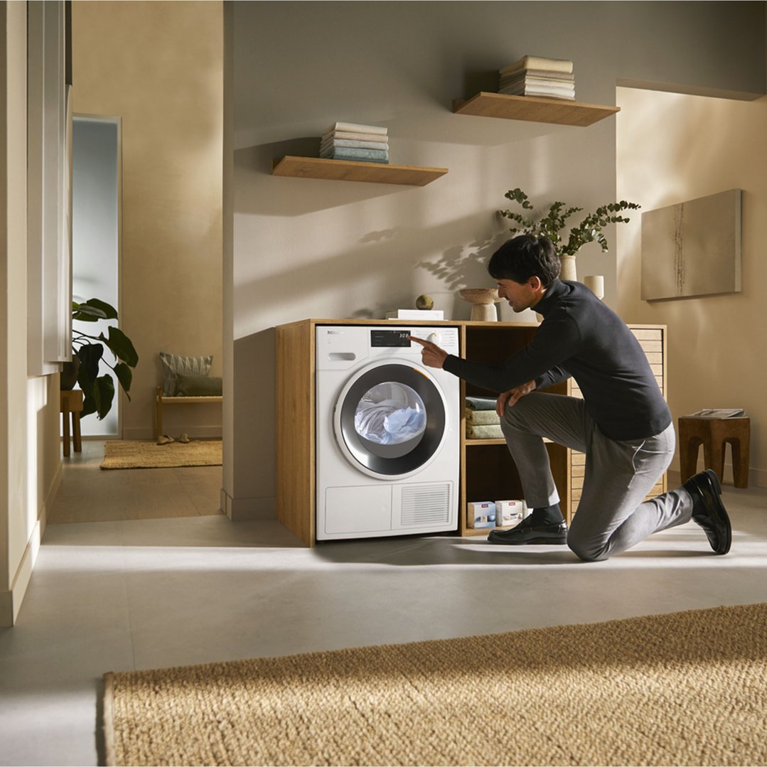 Man kneeling to operate a Miele heat pump dryer in a modern laundry room