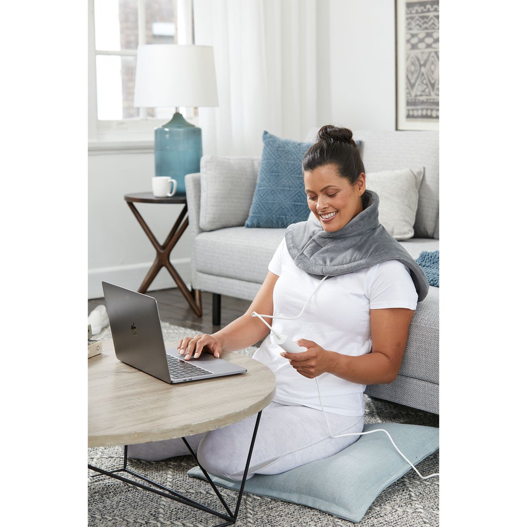 Woman wearing a gray heating pad on her shoulders and neck, using a laptop