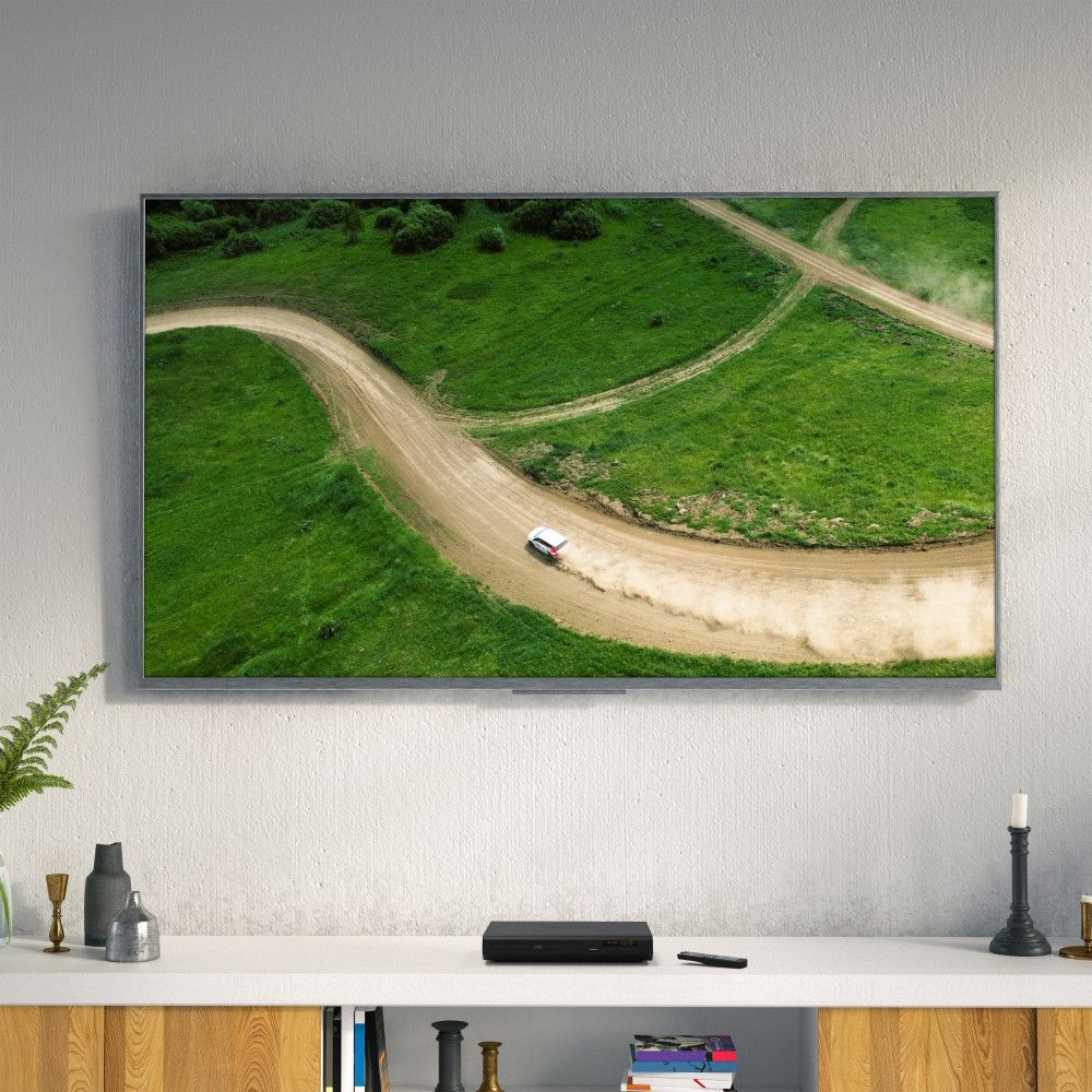 DVD player on a white shelf below a wall-mounted TV showing a rally car