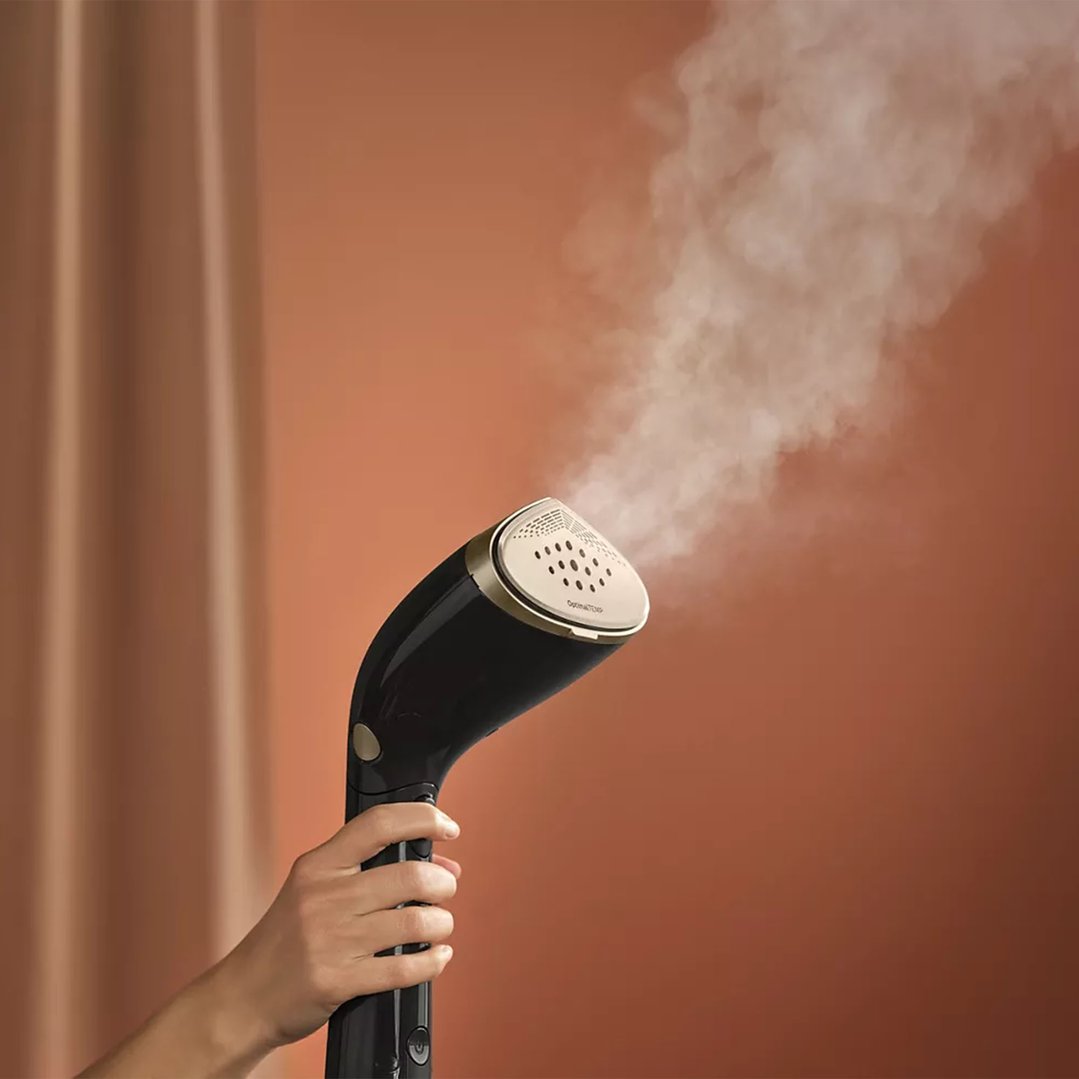 Hand holding a black handheld garment steamer emitting steam against a brown background