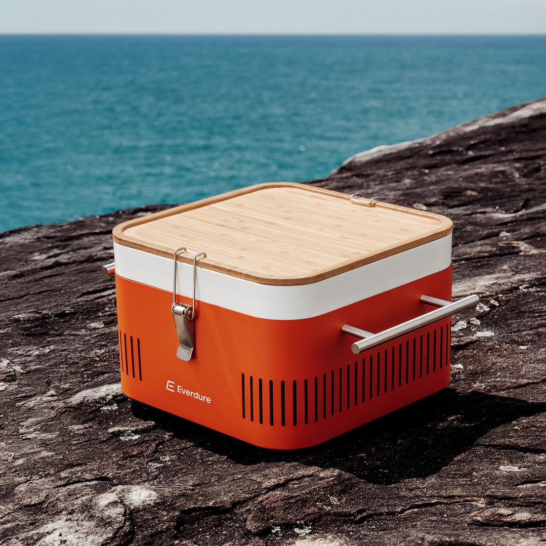 Orange portable charcoal BBQ with a wooden lid on a rock with the ocean in the background