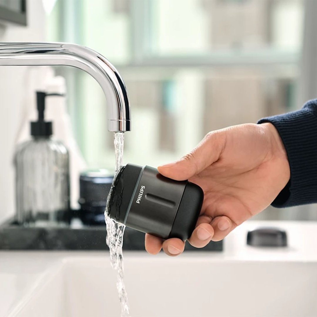Hand holding a Philips electric shaver under running water in a bathroom sink