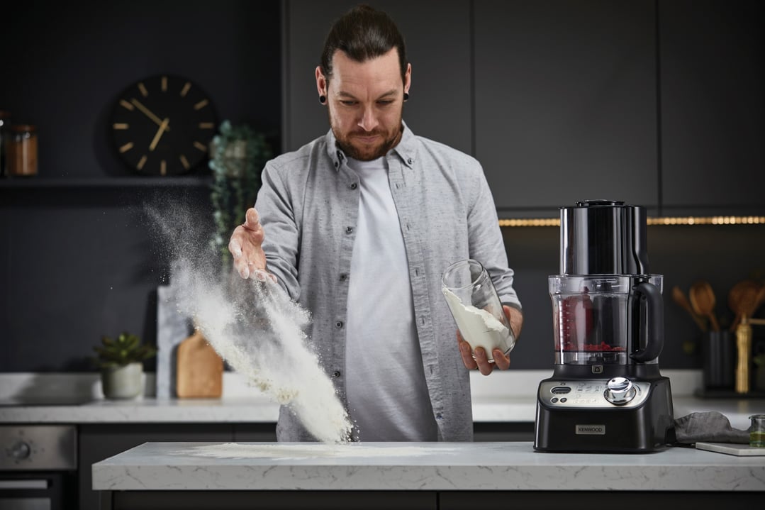 Man sprinkling flour on a kitchen counter next to a food processor