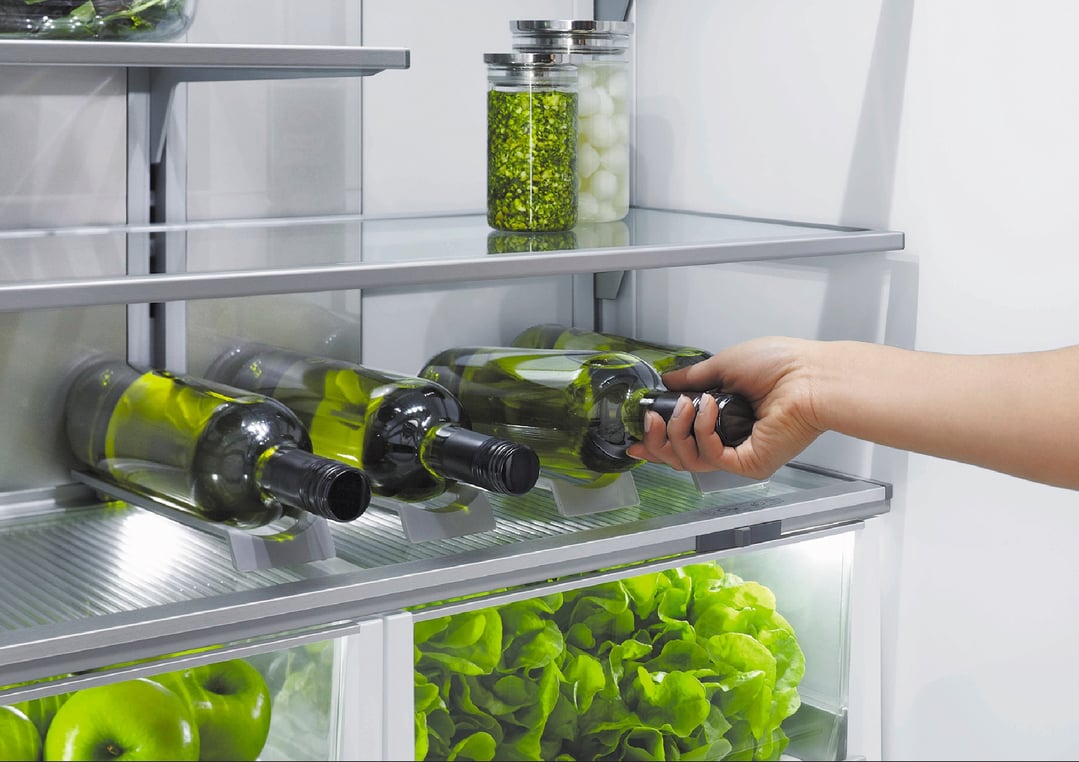 Hand placing a bottle of wine into a refrigerator with fresh produce
