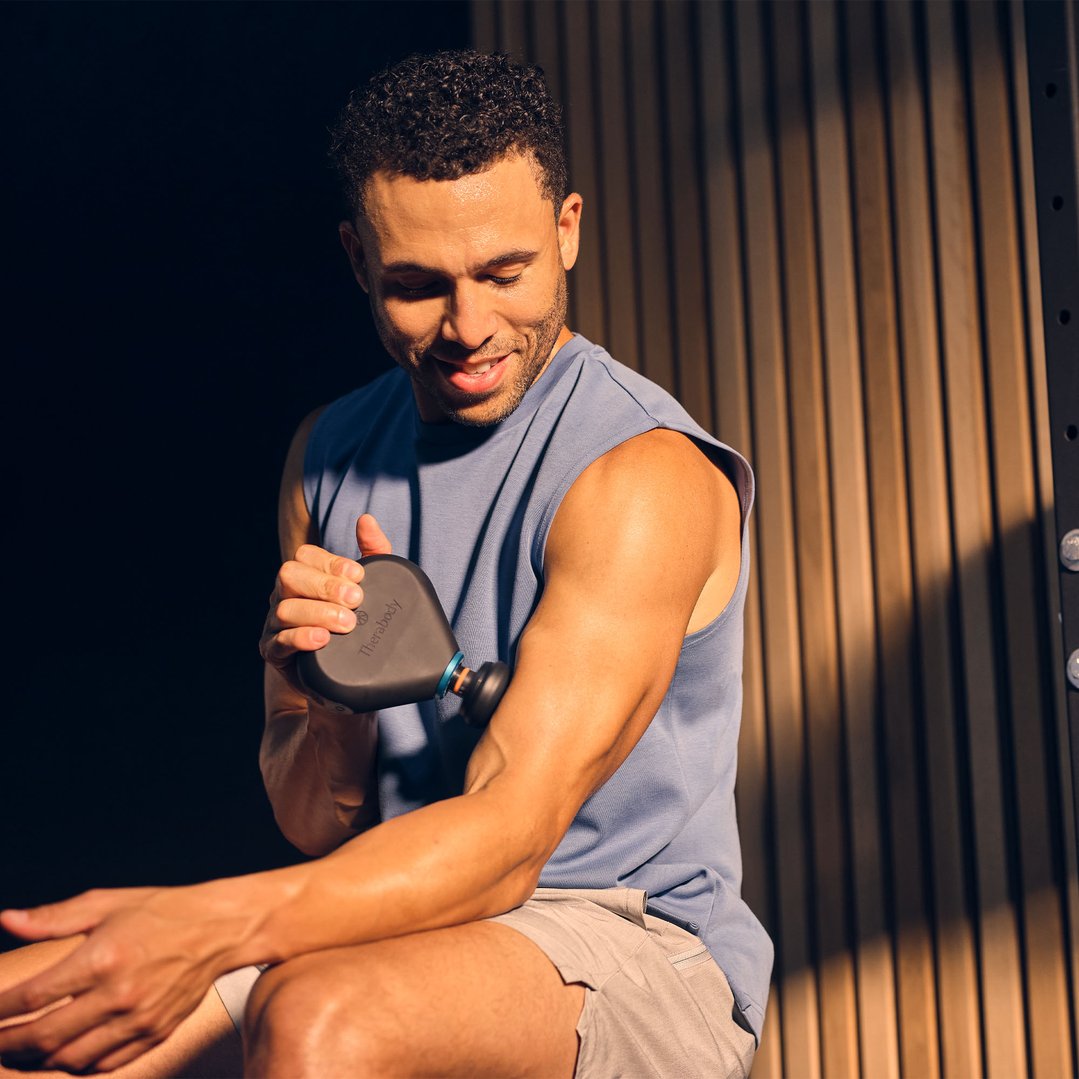 Man using a Theragun Mini Plus massager on his bicep in a gym setting