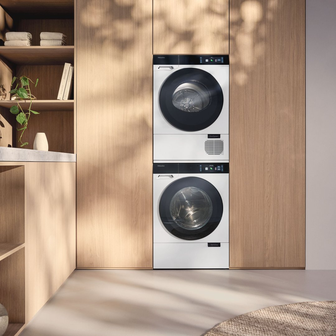 Two Miele washing machines stacked in a wooden laundry room with shelves