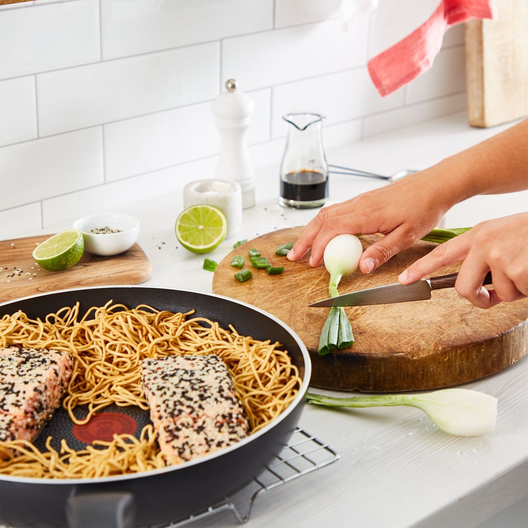 Hands chopping spring onions next to a frypan with noodles and salmon
