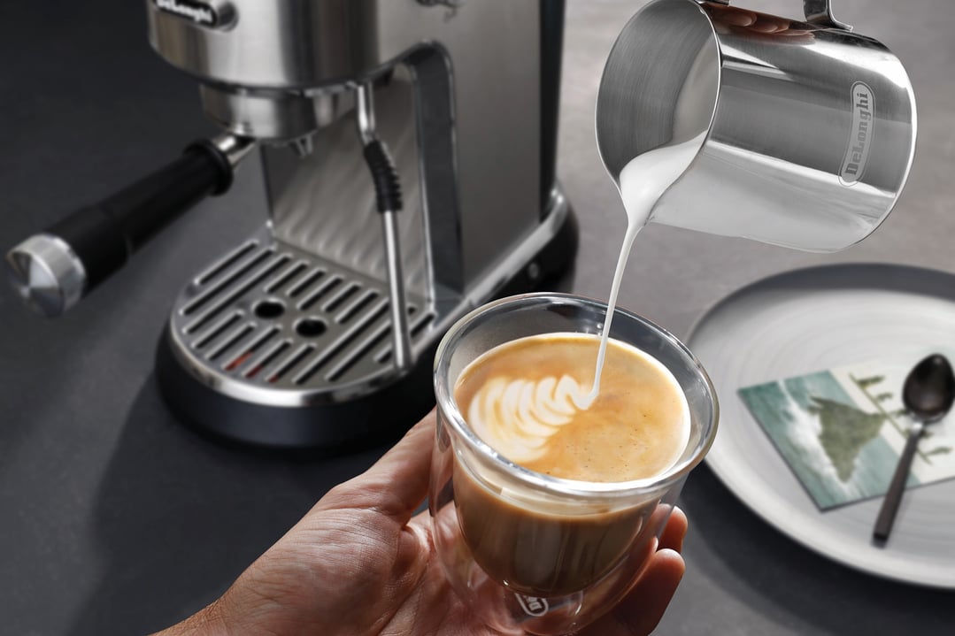Hand pouring frothed milk from a jug into a coffee cup with a coffee machine