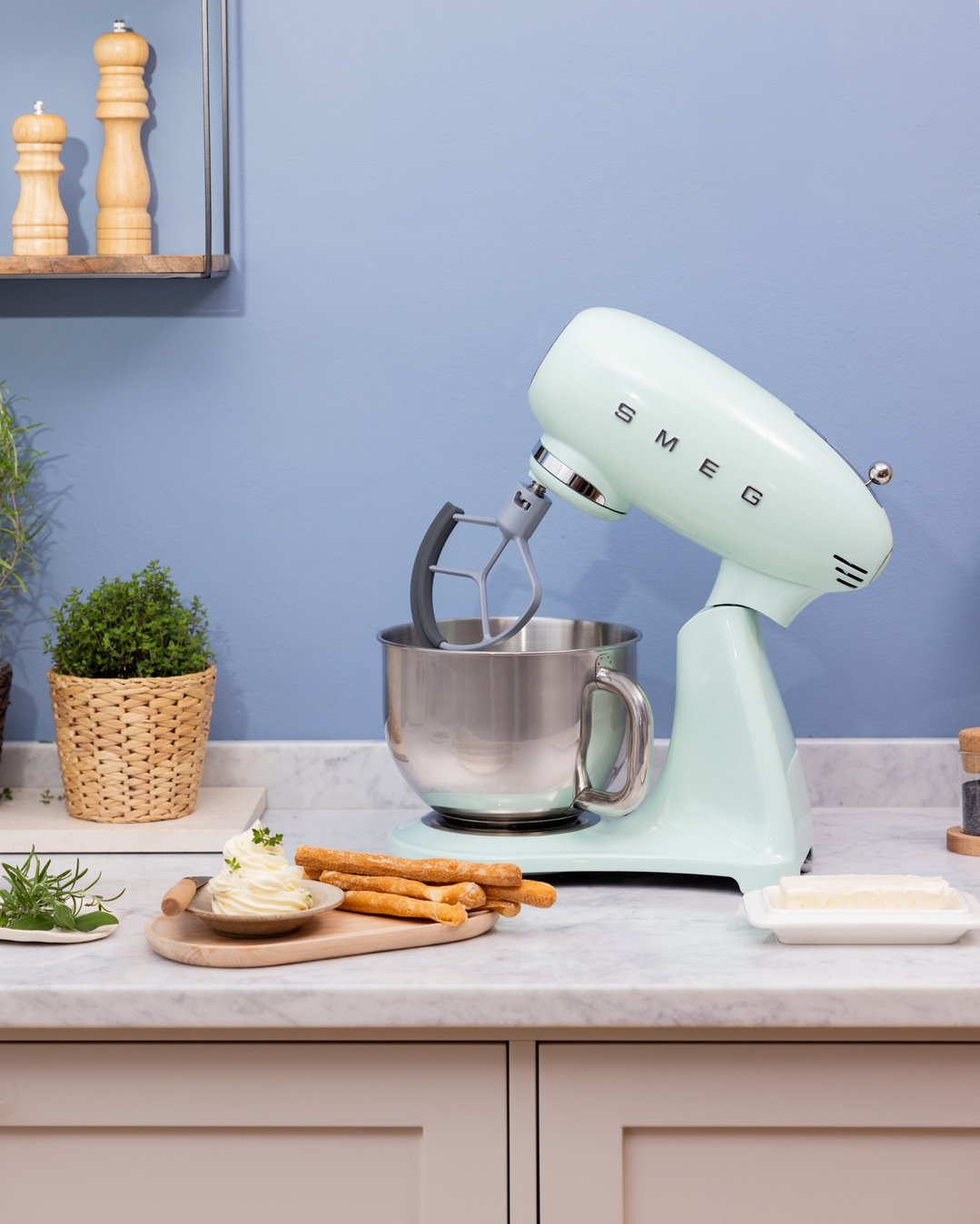Pastel green Smeg stand mixer on a kitchen counter with herbs and breadsticks