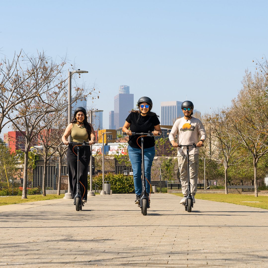 Three people riding electric scooters on a paved path in a park