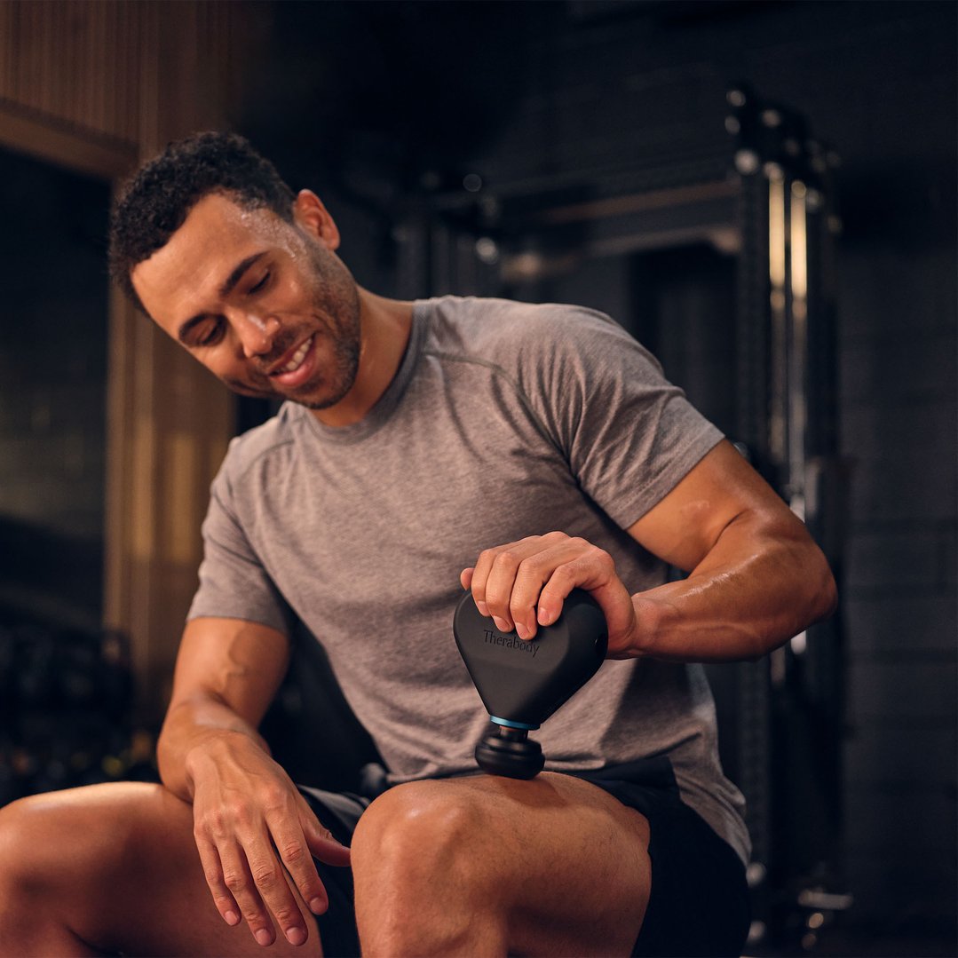 Man using a Theragun Mini Plus massage gun on his leg in a gym