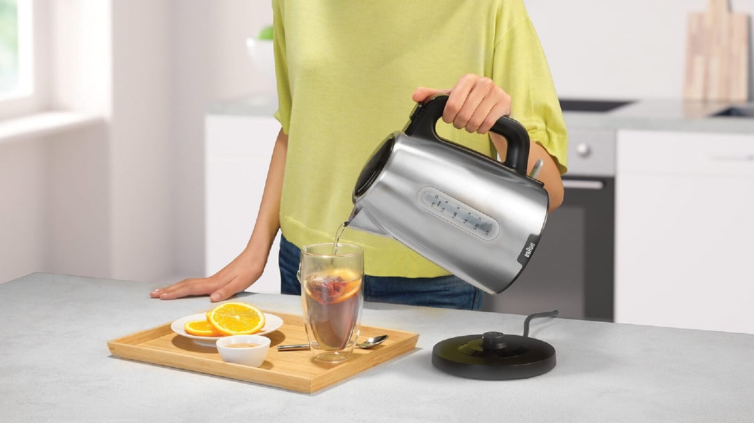 Person pouring hot water from a Braun kettle into a glass on a kitchen counter