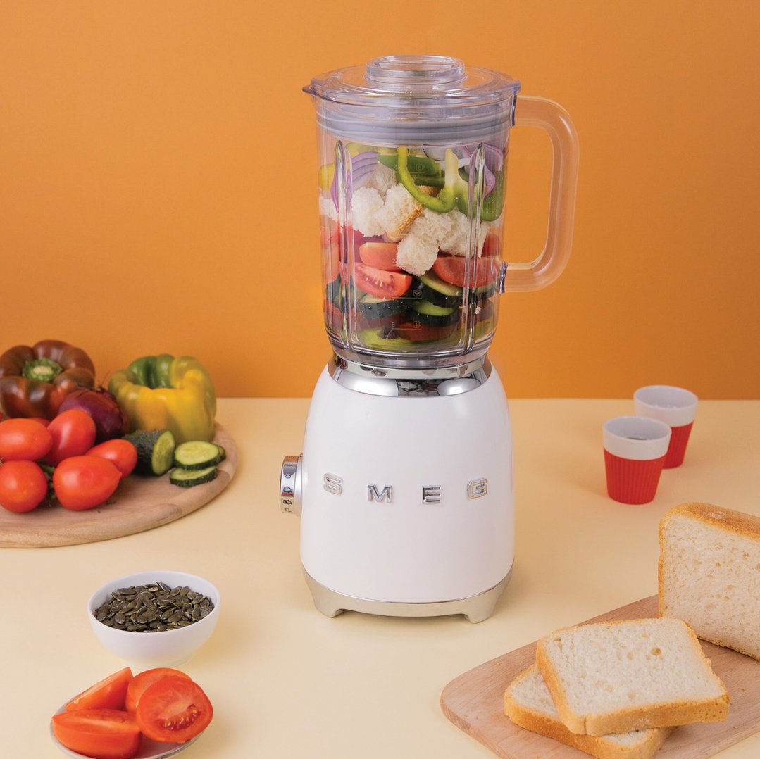 White SMEG blender with vegetables and bread on a light yellow surface