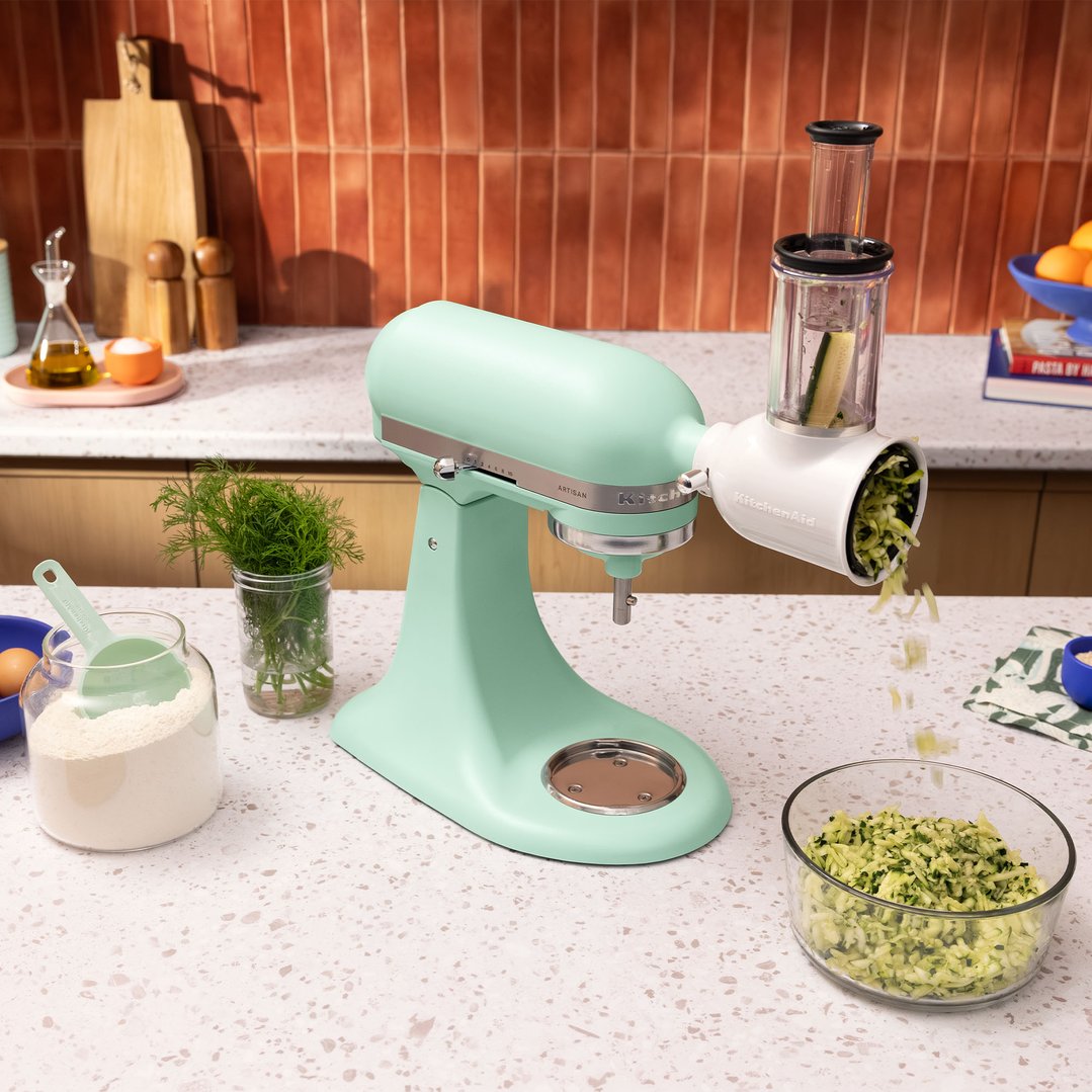 Spearmint KitchenAid stand mixer with a grater attachment shredding zucchini on a kitchen counter