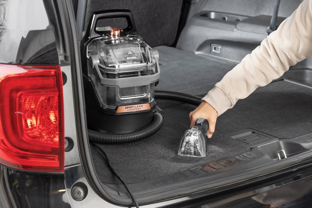Hand using a BISSELL SpotClean HydroSteam vacuum cleaner in the trunk of a car