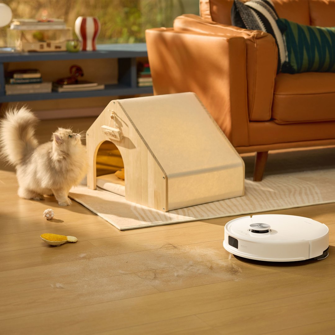 A robotic vacuum cleaner on a wooden floor with pet hair, a cat, and a pet house
