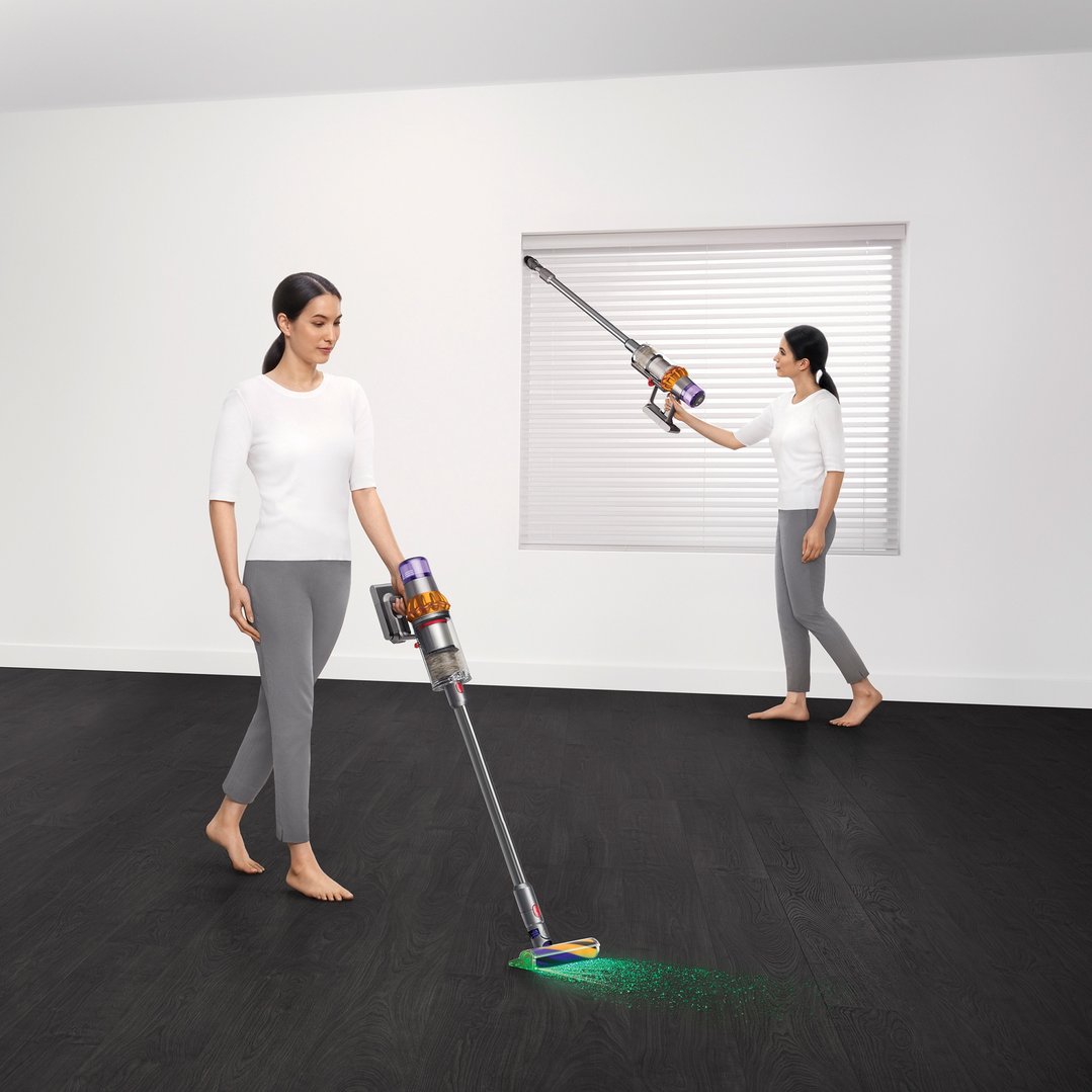 Two women using Dyson handstick vacuums to clean a room with dark flooring