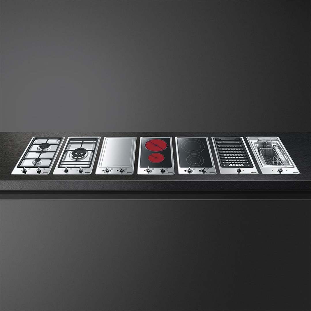 Seven different cooktops and cooking modules arranged on a dark countertop