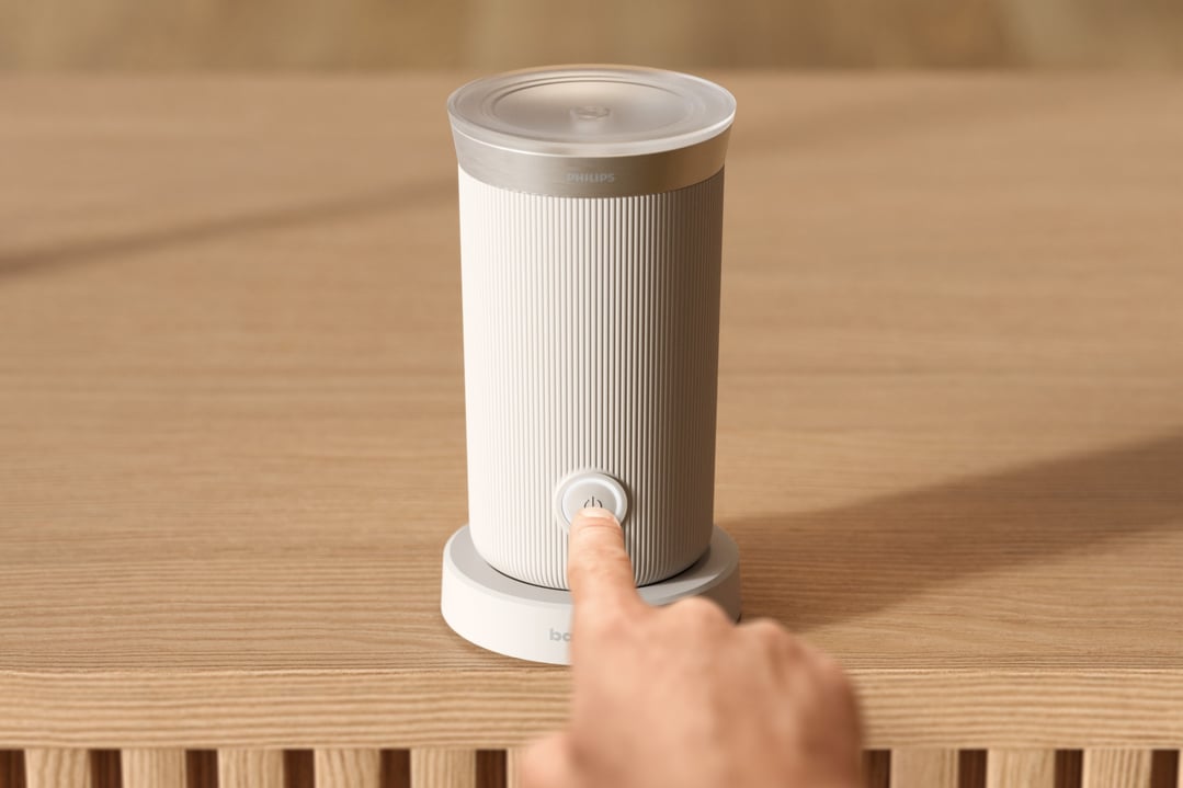 Hand pressing the power button on a Philips Baristina Milk Frother on a wooden surface