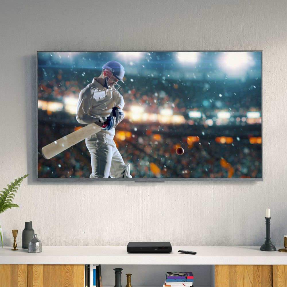 DVD player on a white shelf below a TV showing a cricket match