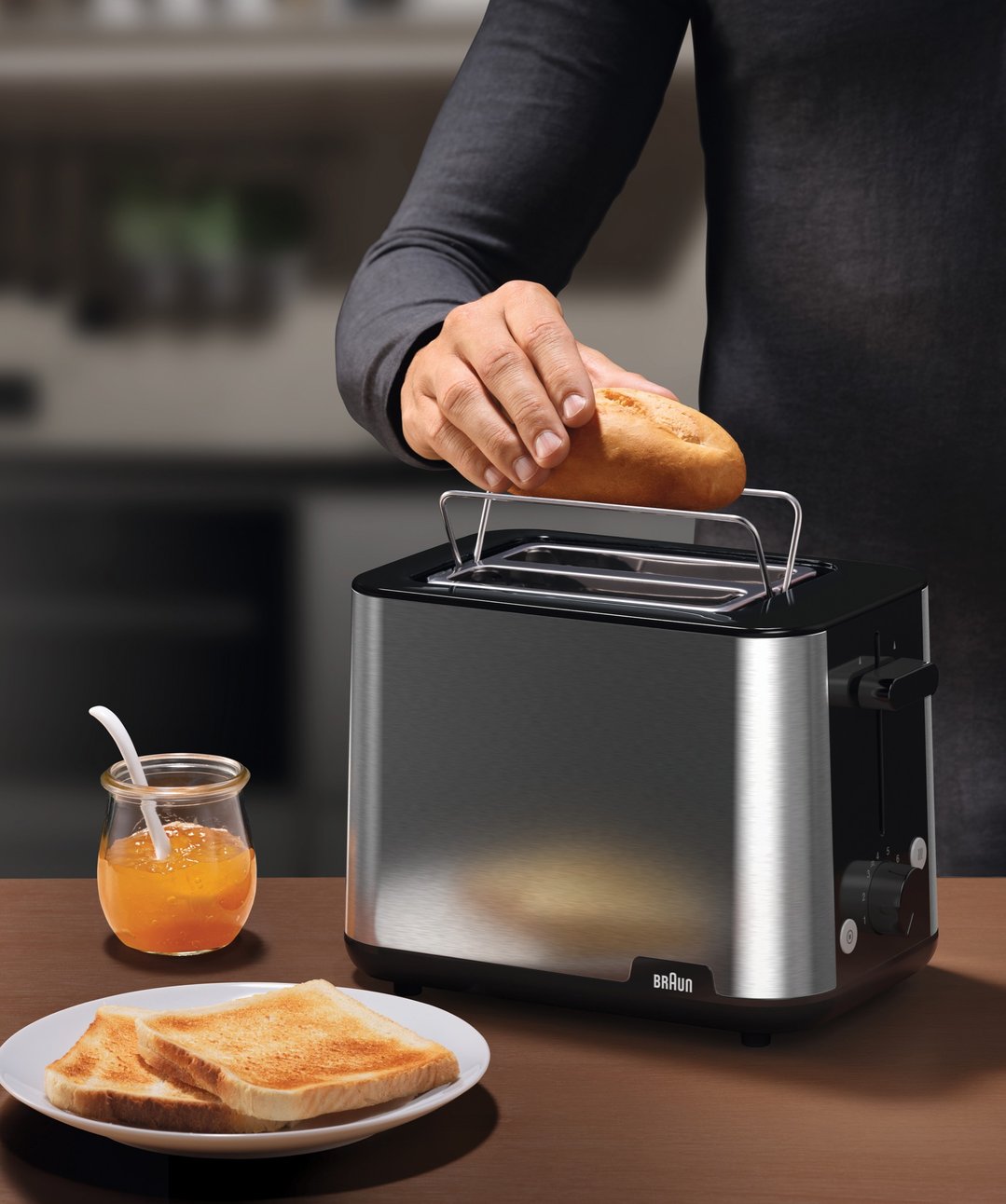 Hand placing bread roll into Braun toaster with toast and jam on a table