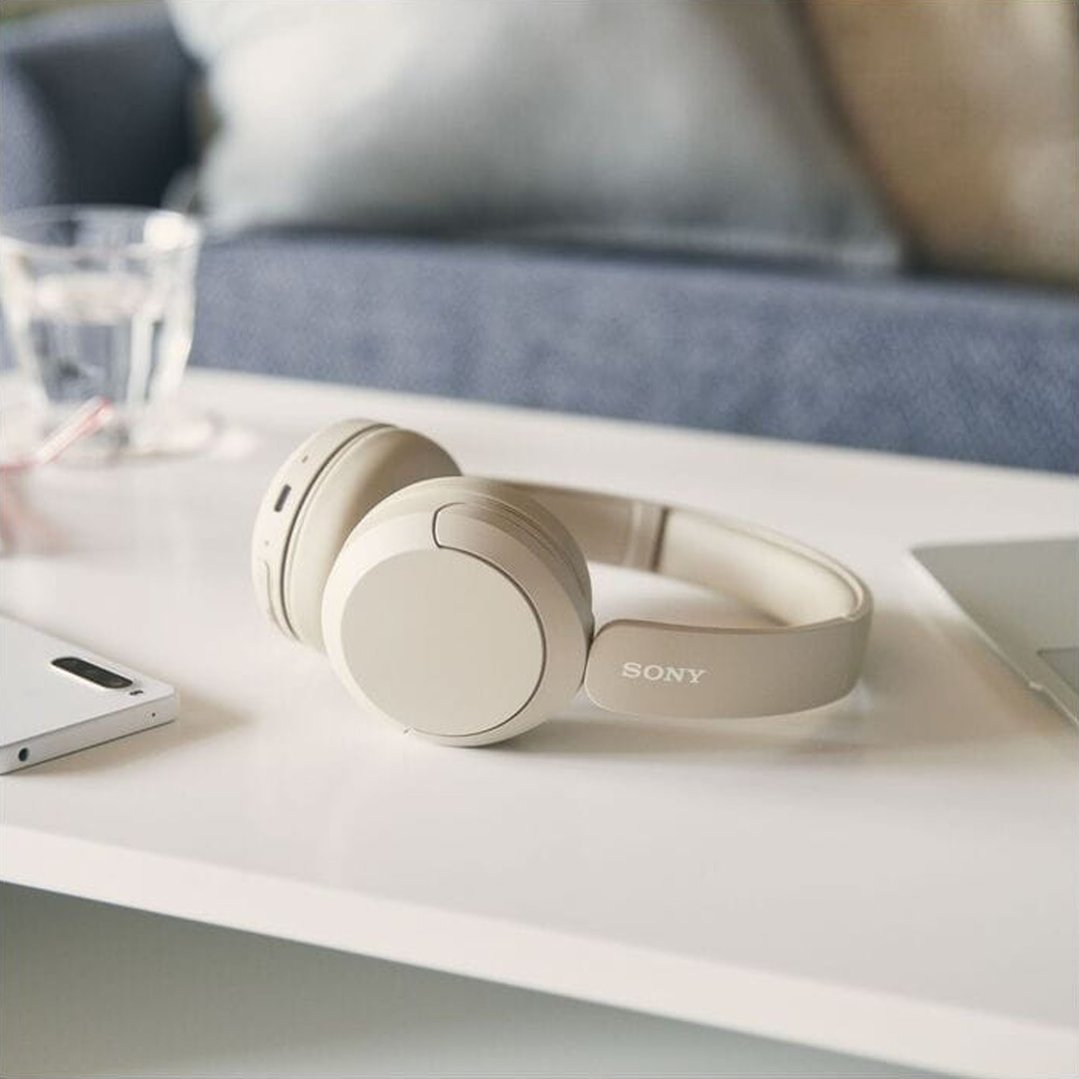 Beige Sony wireless headphones on a white table with a phone and glass