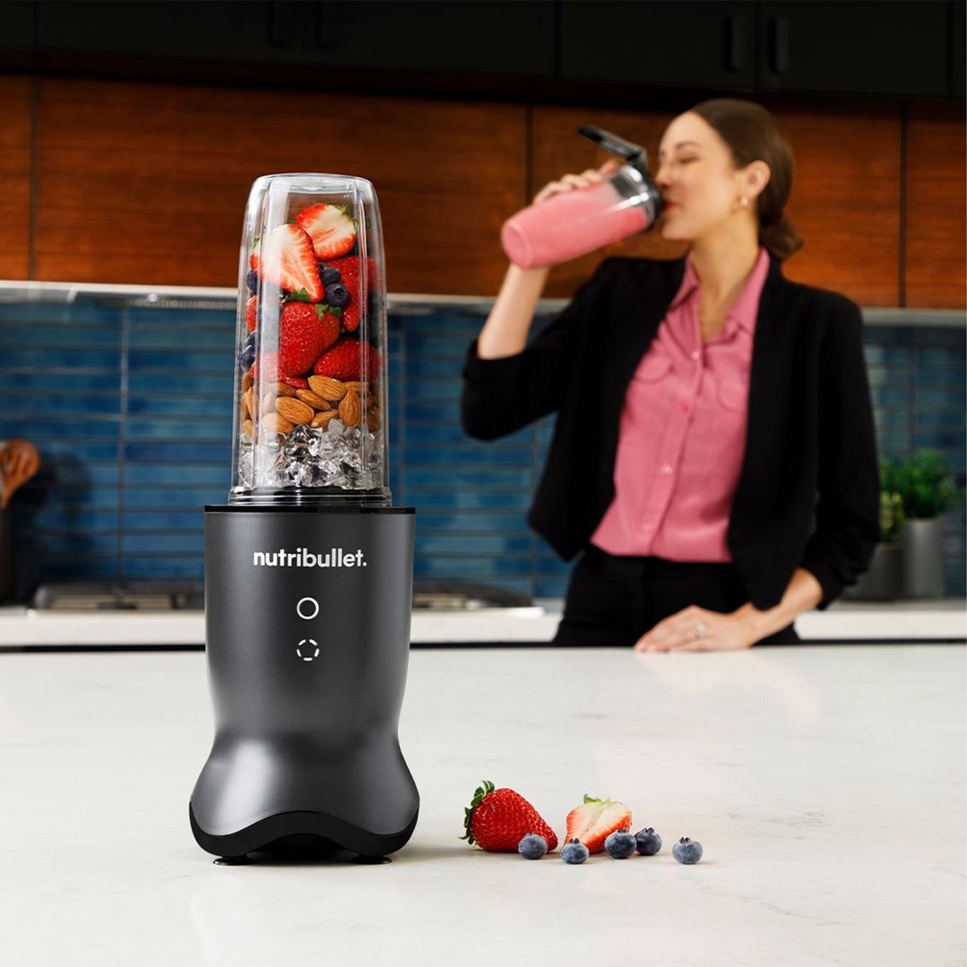 Woman drinking a smoothie while a Nutribullet blender with ingredients sits on a counter