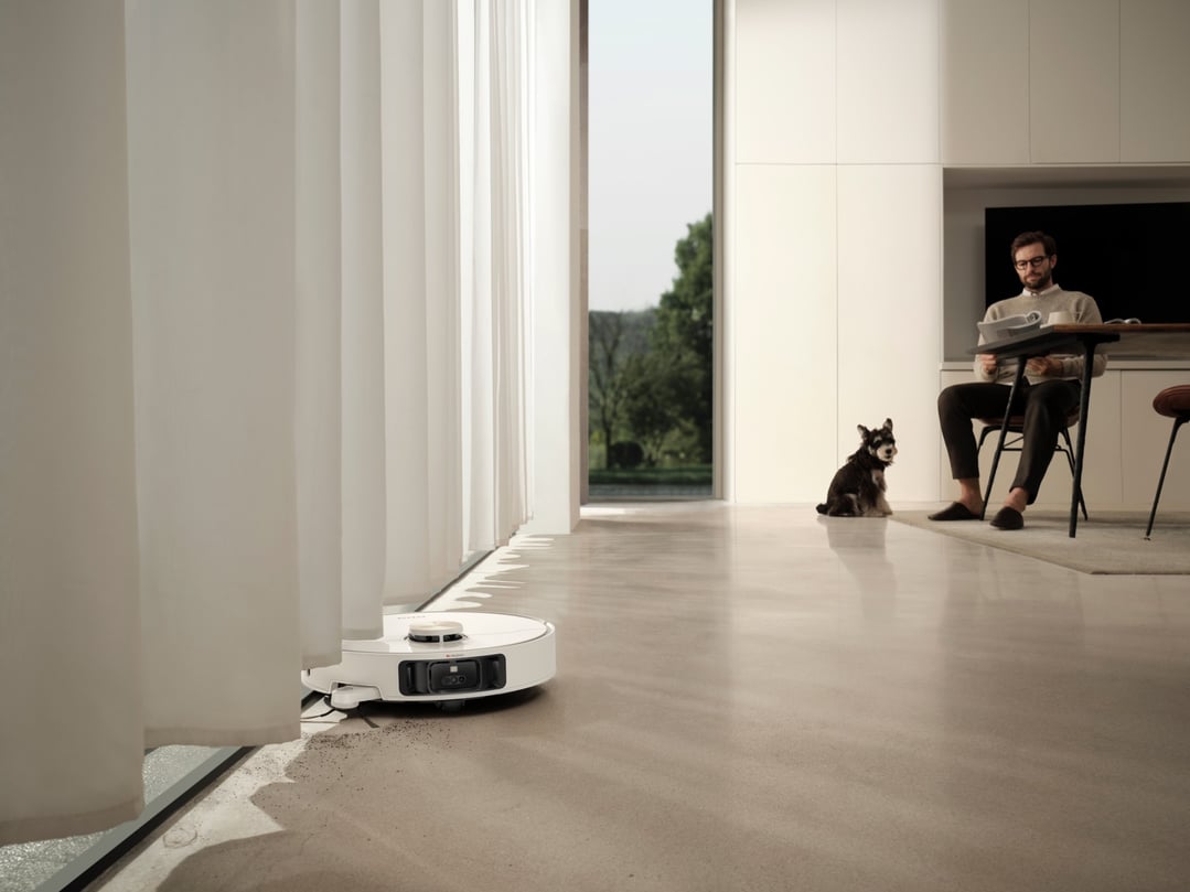 White robot vacuum and mop cleaning floor next to window with man and dog