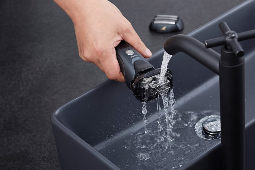 Hand holding a Panasonic shaver under running water in a dark grey sink