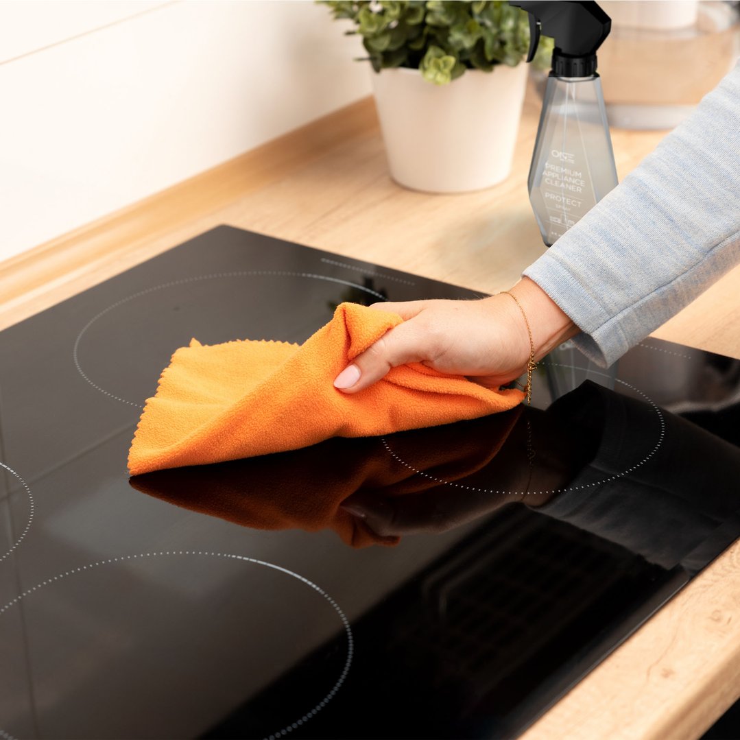 Hand cleaning a black cooktop with an orange cloth and appliance cleaner