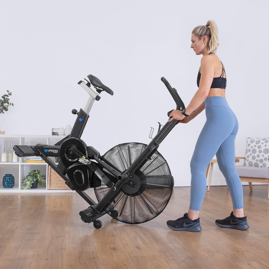 Woman in athletic wear moving a black exercise air bike on a wooden floor