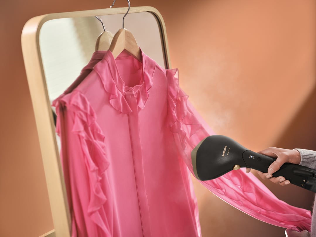 Handheld garment steamer being used on a pink ruffled shirt hanging on a mirror