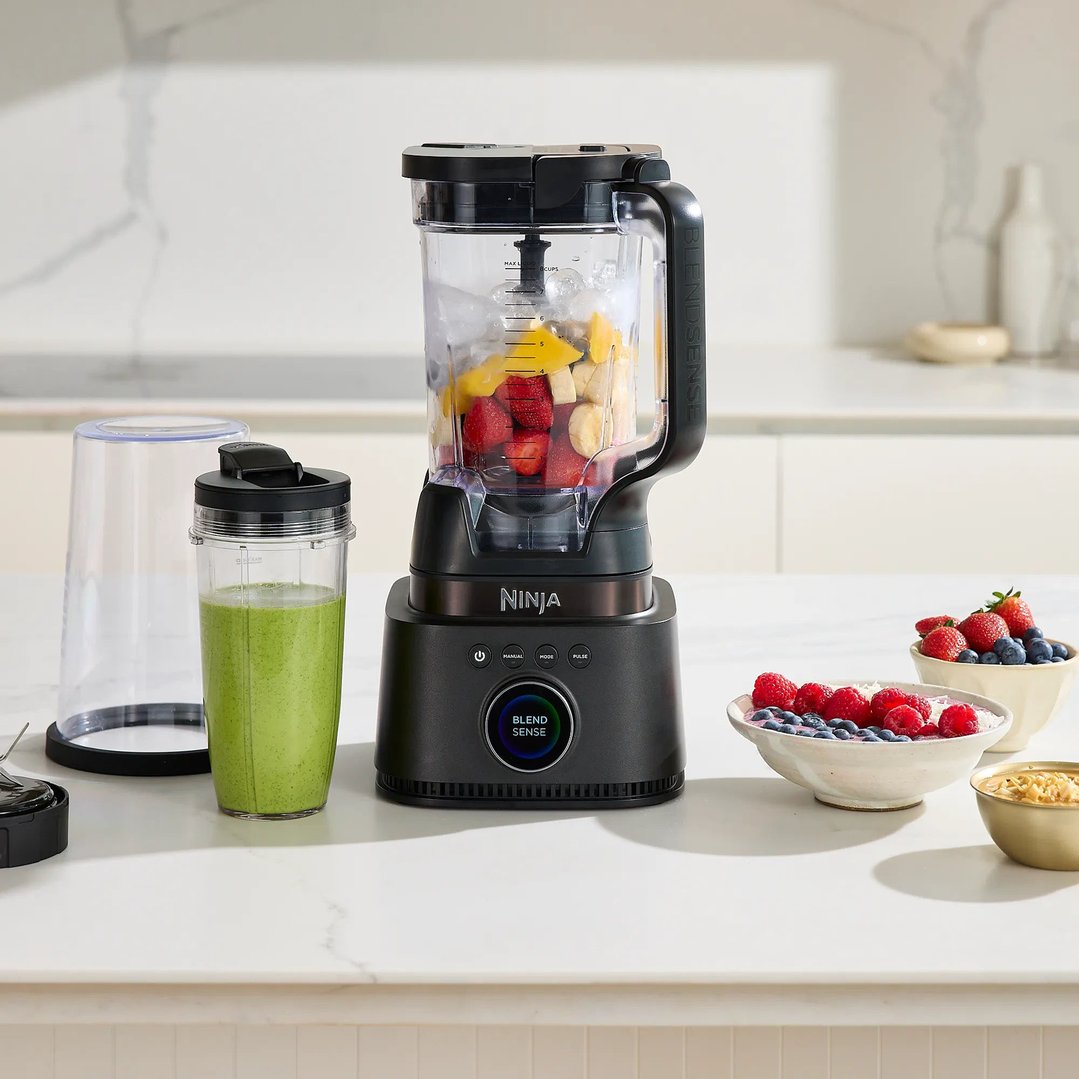 Ninja blender with fruit and ice in pitcher, green smoothie in cup on counter
