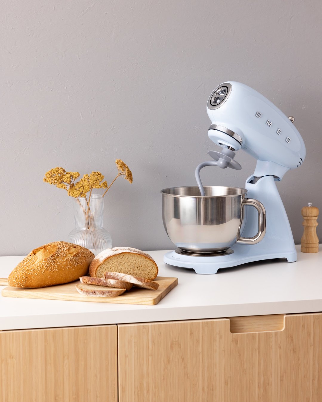 A pastel blue SMEG stand mixer on a kitchen counter with bread and dried flowers